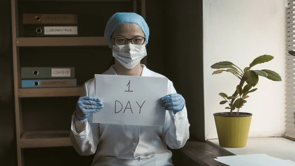 Femeie doctor care deține hârtie goală cu inscripția O zi, înainte de sfârșitul carantinei. Femeia asiatică în uniformă albă stând lângă fereastră. Conceptul numărătoarea inversă. Imagine tonată — Fotografie, imagine de stoc