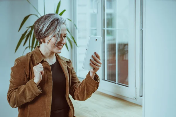 Mooie volwassen vrouw communiceert online op digitale tablet. Zilverharige vrouw glimlacht terwijl ze bij het raam staat. Getinte afbeelding — Stockfoto