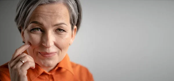 Encantadora mujer de pelo gris sonríe tocando la mejilla con el dedo. Enfoque selectivo en la cara femenina con pequeñas arrugas. Cierre el retrato. Concepto de cuidado de la piel madura. Horizontal espacio de copia witn en blanco para texto —  Fotos de Stock