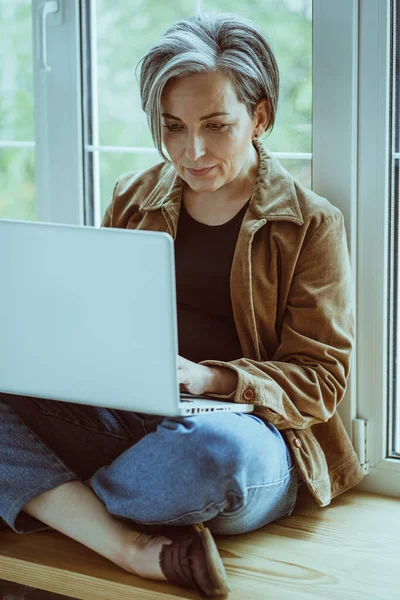 Wanita cantik yang dewasa bekerja di komputer laptop duduk di ambang pintu sambil bersandar ke jendela. Konsep freelance. Citra cetak — Stok Foto