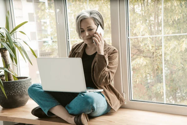 Charmante vrouw die aan de telefoon praat terwijl ze aan de laptop werkt. Gray-haired oude vrouw zit gekruiste benen op brede vensterbank, leunend haar rug tegen het raam. Getinte afbeelding — Stockfoto