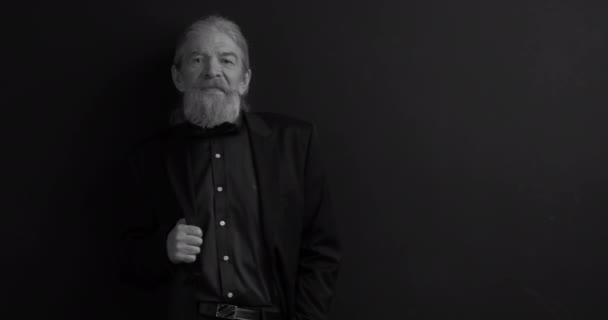 Old business man with long beard in dark business clothes with one hand in chest pocket looking at camera in semi-lit room on black background. Black and white portrait. Waist portrait. Prores 422 — Stock Video