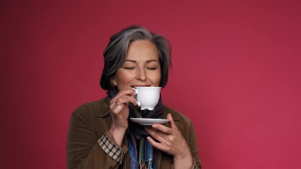 Charmante reife Frau genießt Tasse Kaffee mit geschlossenen Augen. Glückliche grauhaarige Dame trinkt Kaffee und posiert auf rosa Hintergrund im Studio. Nahaufnahme Porträt mit Textfeld rechts. Prores 422 — Stockvideo