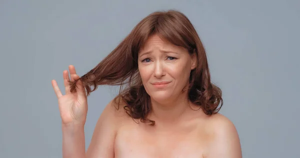 不快乐的漂亮女人拿着头发端头看着摄像机.漂亮的裸体成熟女人在灰色的背上。头发护理概念 — 图库照片