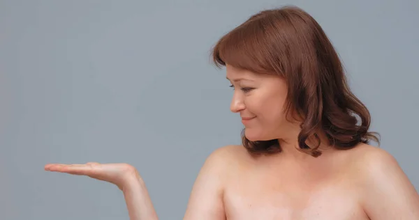 Veselá zralá žena držící otevřenou ruku pro vizualizaci péče o pleť. Usmívající se žena inzerovat anti-age krém izolované na šedém pozadí — Stock fotografie