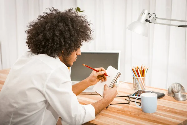 Vista laterale di uomo shaggy prendere appunti nel suo taccuino di carta o gratta e vinci seduto al tavolo di legno con computer e tazza di bevanda calda su di esso. Concetto problemi di memoria. Immagine colorata — Foto Stock