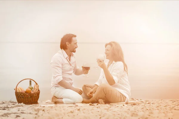 Woman and man drink wine having picnic by water. Two smiling people look each other sitting at sand beach with basket of food next to them. Tinted image — Stock Photo, Image