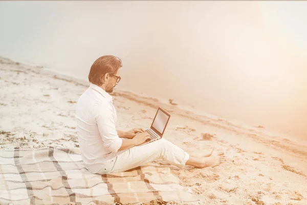 Kopierer arbeitet mit Laptop am Meer. Seitenansicht eines smarten kaukasischen Geschäftsmannes, der Computer benutzt, während er auf einer Decke am Meer sitzt. Getöntes Bild — Stockfoto