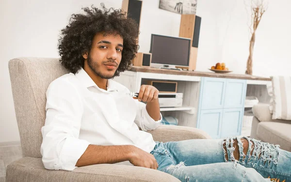 Ung stilig kille rökning av elektroniska eller e-cigarett medan du sitter på fåtölj på modern heminredning bakgrund. Tonad bild — Stockfoto