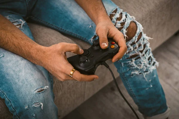 Αρσενικά χέρια κρατώντας χειριστήριο. Μοντέρνος νεαρός με τζιν περνάει χρόνο παίζοντας ηλεκτρονικά παιχνίδια. Η έννοια του τρόπου ζωής. Κοντινό πλάνο. Έγχρωμη εικόνα — Φωτογραφία Αρχείου