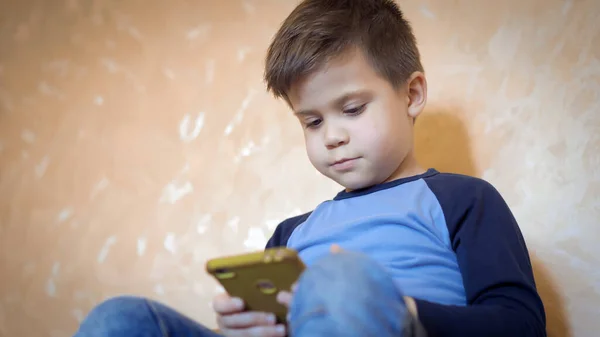 El niño pasa tiempo usando el teléfono sentado en el sofá. Niño jugando o aprendiendo por teléfono móvil en casa. Primer plano. — Foto de Stock