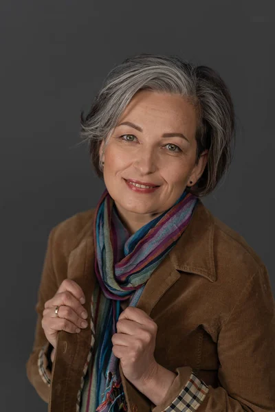 Encantadora mujer madura con ropa casual sonríe fácilmente ajustando el cuello de su chaqueta. Retrato de dama caucásica en chaqueta fustiana marrón y bufanda colorida posando coqueta sobre fondo gris —  Fotos de Stock