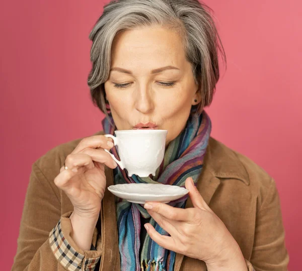 Концепція часу кави. Чарівна кавказька сірувата жінка п'є каву в білій чашці на рожевому фоні в студії. Постріл. — стокове фото