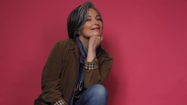 Mujer bonita con el pelo gris sonriendo en la cámara con la mano en la cadera y la mano sosteniendo la cabeza posando para el fotógrafo en el estudio sobre un fondo rosa en ropa casual camisa negra y jeans. Prores 422 — Vídeos de Stock