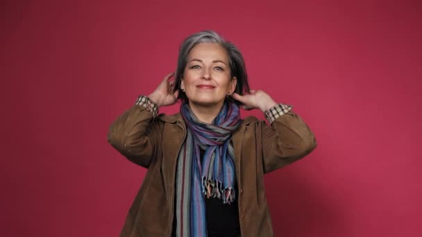 Gray-haired mature woman fixing her hair doing hairstyle. Creative Caucasian woman wearing casual clothes posing in studio on pink background. Prores 422 — Stock Video