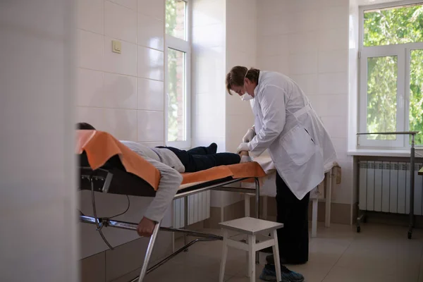 Врач-хирург делает лечение или операцию человеку, лежащему на диване в медицинском кабинете. Городская больница Май, 2020, Бровары, Украина — стоковое фото