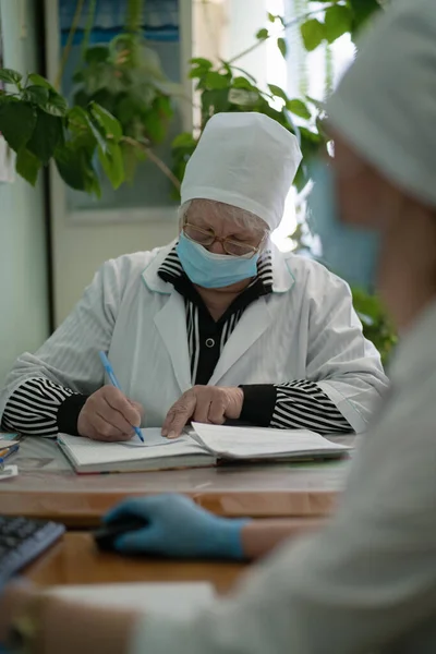 Kvinnlig GP eller sjuksköterska i mask gör anteckningar på medicinska kort. Läkare som skriver i patientkort. Stadssjukhuset. Maj 2020, Brovary, Ukraina — Stockfoto