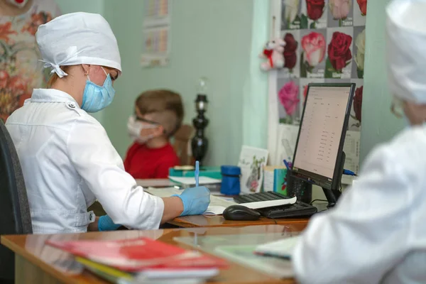 Доктор робить нотатки на картці маленької пацієнтки після обстеження, запису призначень і результатів. Маска жінка і хлопчик через спалах вірусу і пандемію. Міська лікарня. Травень 2020, Броварі, Україна — стокове фото