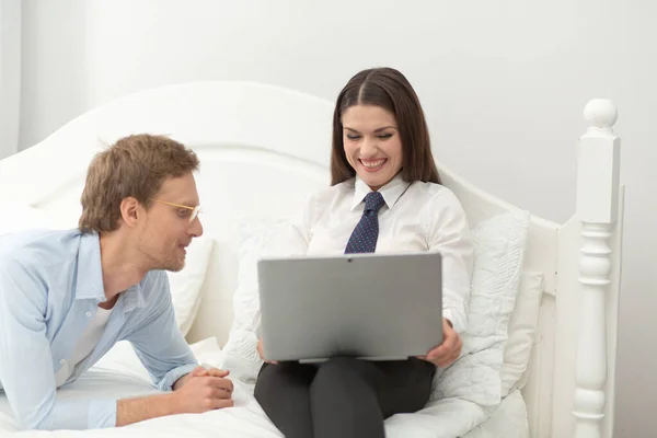 Affärskvinna använder dator med pojkvännen bredvid sig. Vacker kvinna bär business casual kläder skrattar och visar sin man något på laptop när du ligger på sängen — Stockfoto