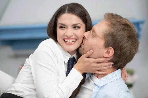 Primo piano di coppia felice a casa. Splendida signora sorridente ampia mentre viene baciata dall'uomo. Uomo che indossa occhiali chiari baciare la sua bella moglie sulla guancia delicatamente mentre lei ride — Foto Stock