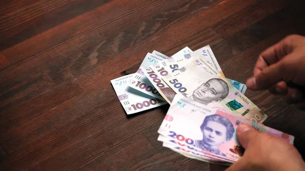 Людина рахує українські гривні гроші. Чоловік тримає пачку нових українських банкнот за різною номіналом на дерев'яному фоні столу. Грошова концепція. Постріл. — стокове фото