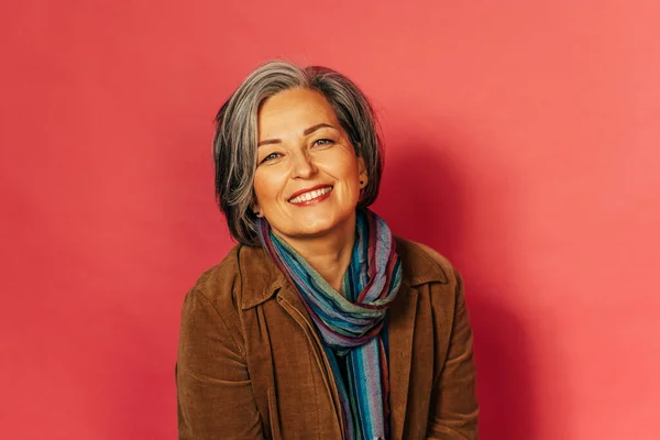 Stylish grey haired woman smiles broadly posing in studio on pink background. Charming good-looking mature lady looks at camera. Glamour concept. Tinted image