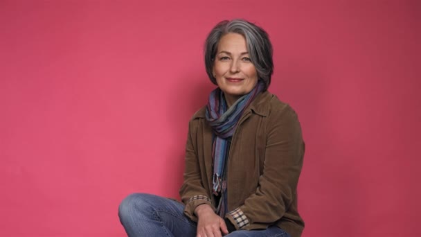 Femme d'affaires indépendante forte avec les cheveux gris souriant à la caméra posant pour photographe en studio sur un fond rose en vêtements décontractés veste brune et jeans. Prores 422 — Video