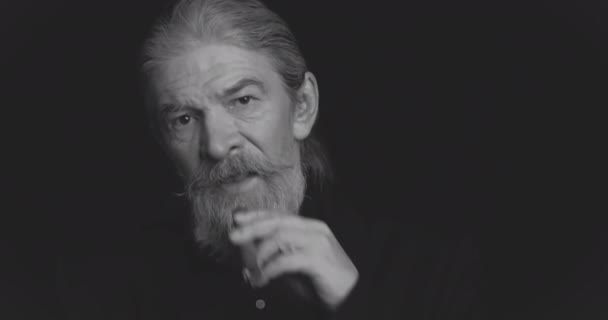 Black and white. Smoking cigar old man with strong facial expressions with long beard in dark business clothes looking at camera in semi-lit room on black. Toned footage. Prores 422 — Stock Video