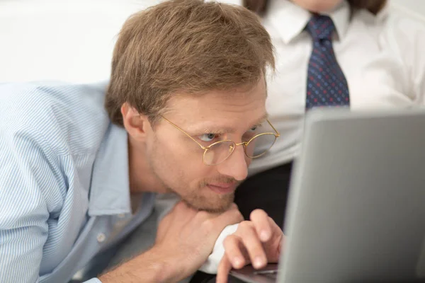 Mannen som tittar på när hans fru jobbar på datorn. Närbild porträtt av man bär runda tunna glasögon på sin flickvän på sängen och titta på hennes arbete på laptop — Stockfoto