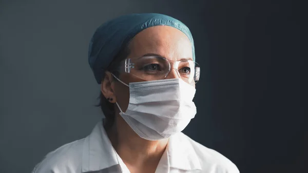 Medicul mascat în ochelari de protecție care se uită la o parte. O femeie caucaziană de vârstă mijlocie în haină albă pe fundal gri închis. Închide portretul. Conceptul de sănătate. Imagine tonată — Fotografie, imagine de stoc