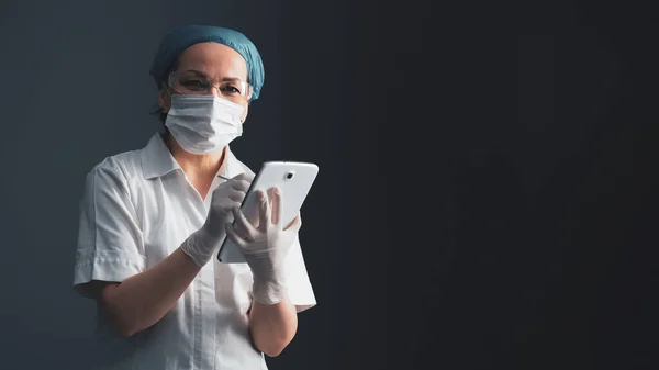 Médica usa comprimido digital. Mulher de casaco médico fica em fundo cinza. Tecnologia moderna e conceito de saúde. Em branco para banner com emty place à direita. Um tiro de perto. Imagem matizada — Fotografia de Stock