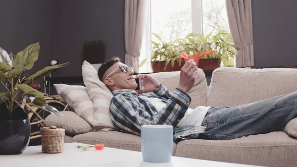Young man talks mobile phone lying on sofa. Smiling guy communicates via cellular holding darts arrow. Rest concept. Tinted image — Stock Photo, Image