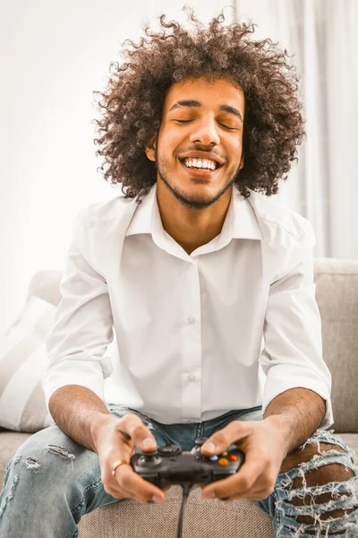 Shaggy gamer closed his eyes with pleasure and laughs cheerfully while playing computer game, pressing buttons of game console. Young man play computer game at home in self isolation — Stock Photo, Image