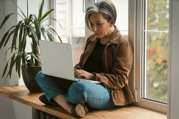 Σοβαρή ώριμη γυναίκα σε casual χρησιμοποιεί φορητό υπολογιστή κάθεται σταυρωμένα πόδια σε ξύλινο περβάζι παράθυρο. Σκυμμένη εικόνα. Έννοια της ελευθερίας — Φωτογραφία Αρχείου