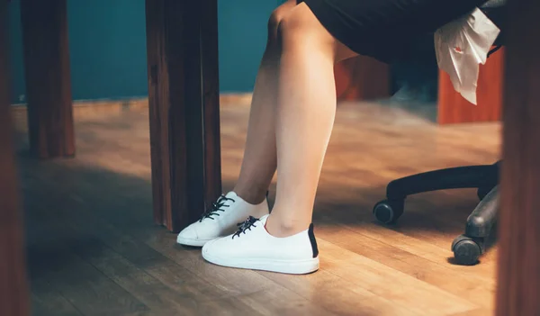 Zakelijke vrouw benen in witte sneakers. Jonge blanke vrouw in comfortabele schoenen werkend op kantoor. Dichtbij schot. Getinte afbeelding — Stockfoto