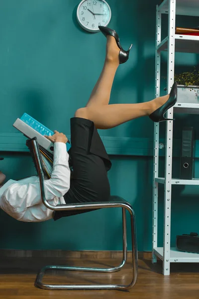 Unternehmerinnen haben Spaß bei der Arbeit. Lustige Frau liegt mit erhobenen Beinen auf einem Bürostuhl und hält Arbeitsmappen in den Händen. Schnittwunden. Getöntes Bild — Stockfoto