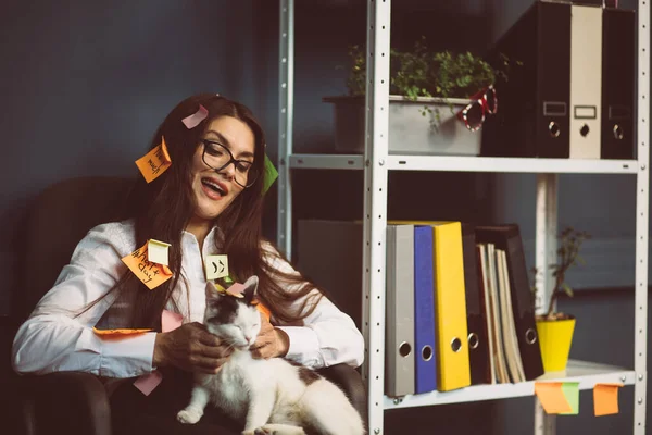 Concept de procrastination. Femme d'affaires folle avec des autocollants de mémoire collés caressant un chat. Fille occupée se détendre dans le bureau. Image teintée — Photo