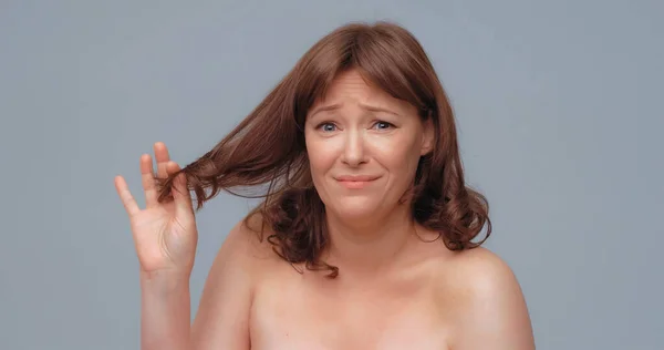 裸露的成熟女人拿着头发端看着相机.不快乐的漂亮女人的头发分叉成灰色背景是有问题的。保健和护理概念 — 图库照片