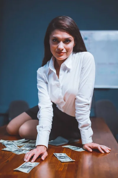 Femme d'affaires de beauté assise sur le bureau avec de l'argent en papier dessus. Jeune fille en tenue de cérémonie et des billets de cent dollars éparpillés sur la table. Image teintée — Photo