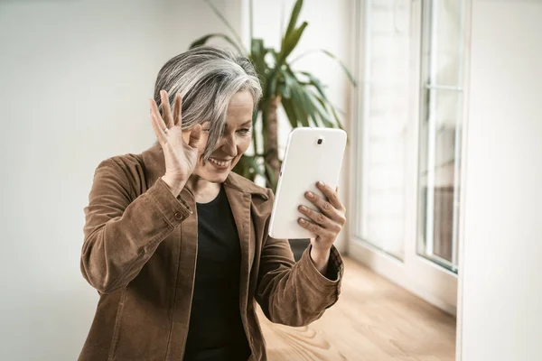 Joyful woman waves her hand using tablet computer for communication with loved ones in internet. Happy aged woman have online conversation on digital tablet. Tinted image — Stock Photo, Image