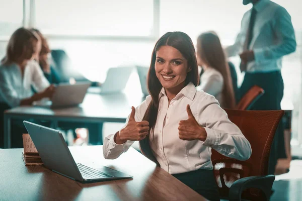 Mulher de negócios mostra os polegares no escritório. Tudo bem gesto de menina sorridente jovem. Grupo de empresários trabalham em fundo turvo. Realização no conceito de negócio. Imagem matizada — Fotografia de Stock