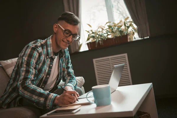 Un joven que trabaja en casa usando tecnologías modernas. Hombre creativo trabajando en el proyecto sentado en la pequeña mesa de café. Concepto independiente. Vista lateral. Ángulo holandés — Foto de Stock