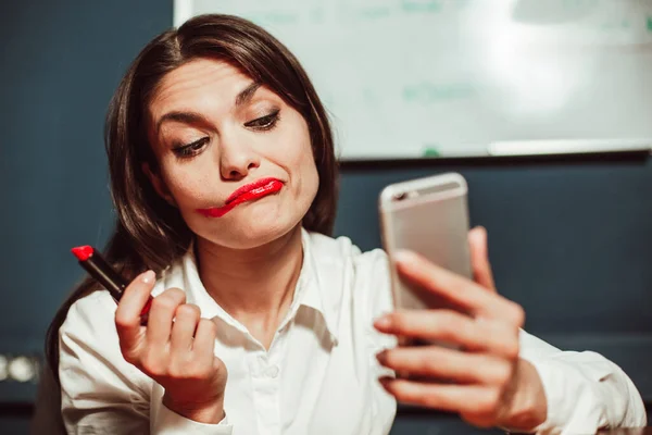 Crazy office manager businesswoman doing makeup with red lipstick at workplace using mobile phone instead of mirror. Tinted image — Stock Photo, Image