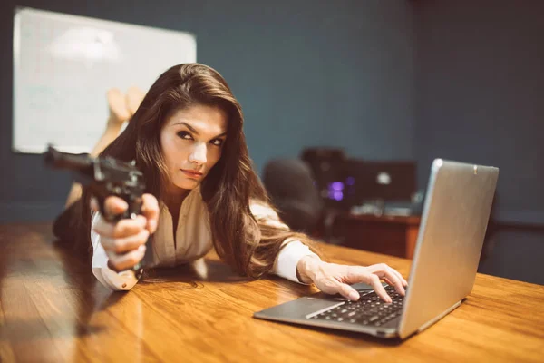 Armada mulher de negócios sexy com pistola obtendo dados do computador. Uma mulher perigosa com uma arma está na mesa do escritório. Imagem matizada — Fotografia de Stock