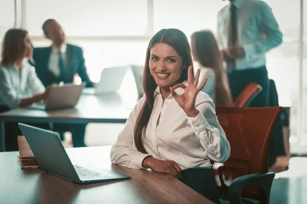 Empresária mostra sinal ok trabalhando no escritório de espaço aberto. Mulher caucasiana feliz com laptop mostrando gesto certo contra seus colegas de trabalho em fundo turvo. Imagem matizada — Fotografia de Stock