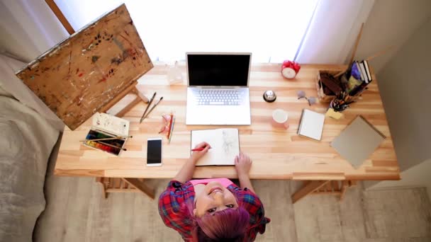 Girl is sitting at the desk. Shes writing in a notebook. On the table is a laptop, writing supplies, paints and brushes. Girl is left-handed. Top View. High quality FullHD footage — Stock Video