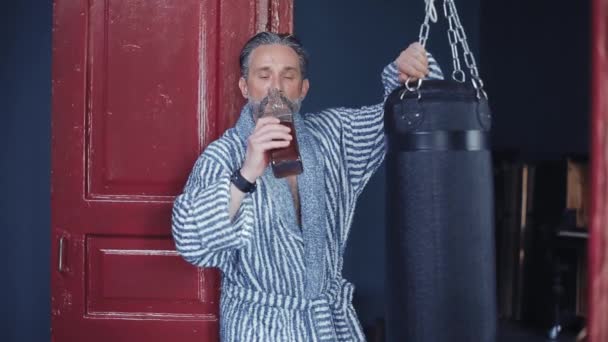 Пьяный бородатый человек игнорирует спорт. Он стоит в халате с бутылкой виски, опираясь на боксерскую грушу. Пьет алкоголь и гладит боксерскую грушу. Высококачественные FullHD кадры — стоковое видео