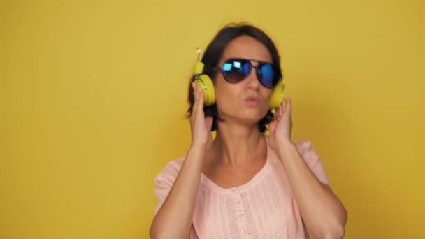 Vrouwen van middelbare leeftijd met bruin haar en blauwe glazen luisteren muziek in gele koptelefoons op de gele achtergrond. Meisje heeft plezier tijdens het luisteren melodie. — Stockvideo