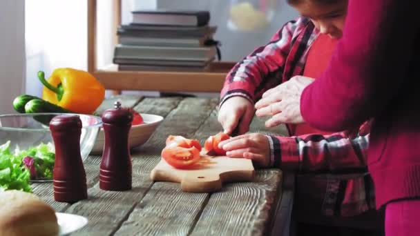 Gammal blond kvinna och ung brunhårig flicka skär tomat tillsammans på köket. Barnbarn och farmor relation. Lycklig familj. Ha kul i köket. Mormor lär barnbarn hur man skär mat. — Stockvideo