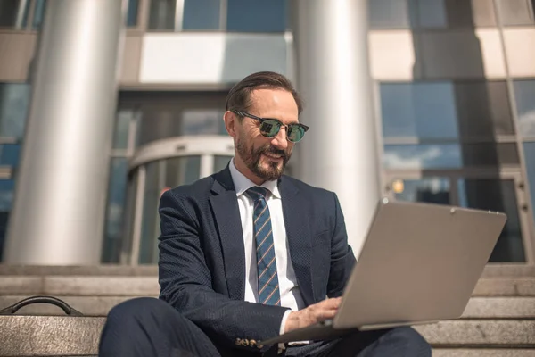 Успішний бізнесмен працює на ноутбуці, сидячи на сходах бізнес-центру. Усміхнений чоловік у діловому костюмі та сонцезахисних окулярах. Високоякісна фотографія — стокове фото